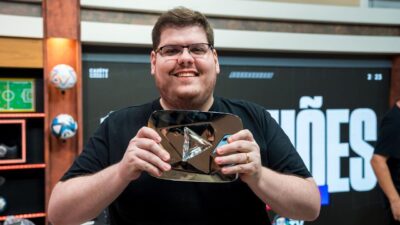 Casimiro Miguel sorri segurando a placa de diamante do YouTube, veste camiseta preta em cenário de estúdio com bolas de futebol e painel ao fundo