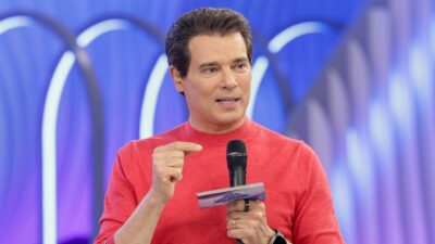 Celso Portiolli veste blusa vermelha, segura microfone preto e fichas do programa Domingo Legal, gesticula com a mão direita em cenário azulado
