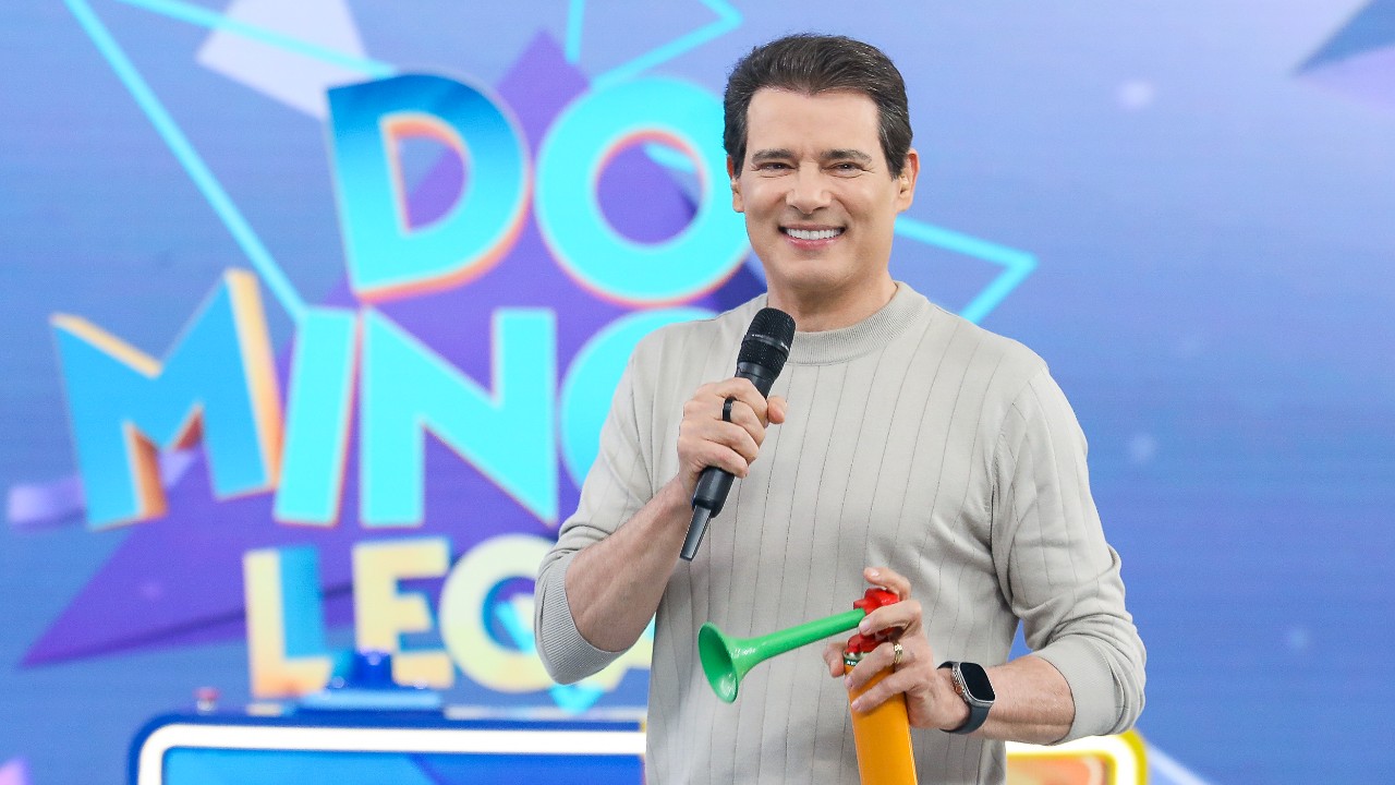 Celso Portiolli sorridente no palco do Domingo Legal, segurando microfone preto e buzina colorida, veste blusa bege de manga longa em cenário azul