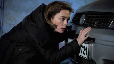 Demet Evgar como Cemre na novela Chamas do Destino está agachada em frente a um carro cinza, usa casaco acolchoado preto e toca o para-choque próximo à placa