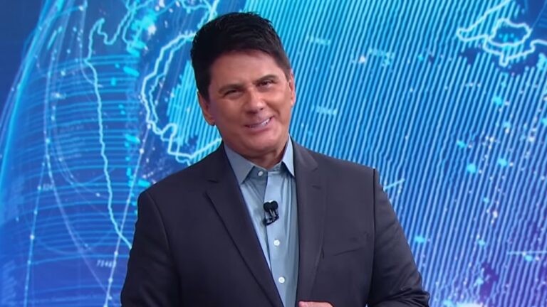 O apresentador César Filho sorri no cenário do SBT Brasil, ele veste paletó escuro sobre camisa azul clara e ao fundo há um mapa mundi tecnológico