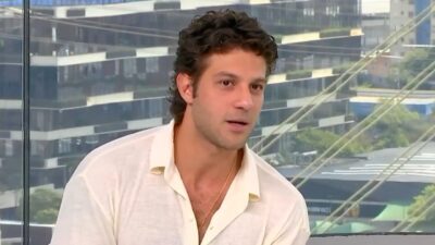 Chay Suede fala durante entrevista em estúdio com janela ao fundo mostrando prédios e ponte na cidade