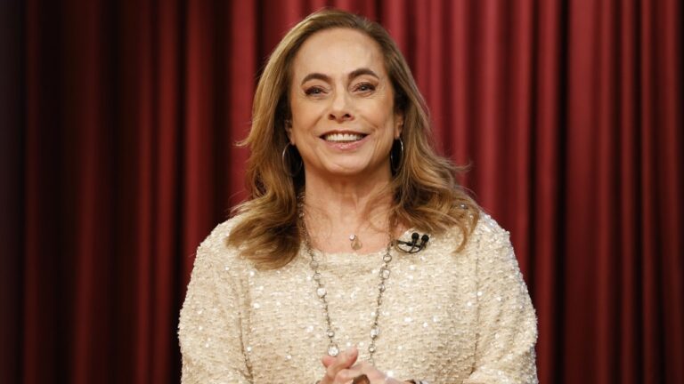 Cissa Guimarães sorri usando blusa bege com brilhos e colar longo, cabelos loiros na altura dos ombros, posicionada à frente de cortina vermelha clara