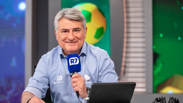 Narrador Cléber Machado sorri e segura microfone azul com logo do R7, veste camisa social azul clara da Record, em estúdio com telas e fundo verde