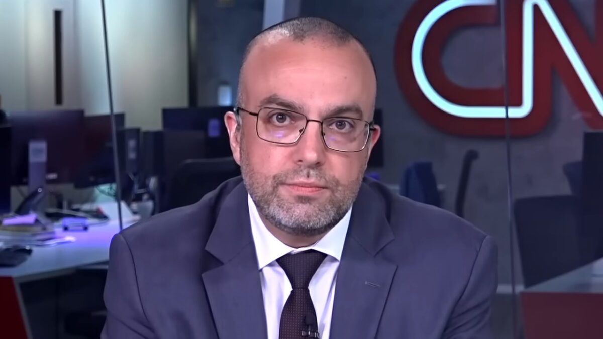 Jornalista Daniel Rittner da CNN Brasil, usa paletó cinza, gravata escura, óculos de grau, possui barba curta, ao fundo logotipo da emissora e redação iluminada
