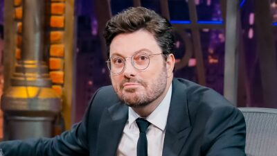 Danilo Gentili usa terno escuro, gravata preta e óculos redondos de grau, mantém expressão séria e está sentado em cenário de estúdio de televisão