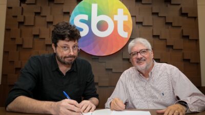 Danilo Gentili de camisa preta assina documento ao lado de Leon Abravanel, que veste camisa listrada, sob o logotipo colorido do SBT em parede de madeira