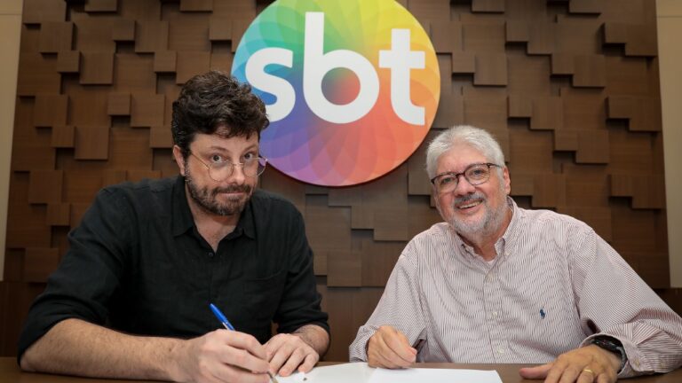 Danilo Gentili de camisa preta assina documento ao lado de Leon Abravanel, que veste camisa listrada, sob o logotipo colorido do SBT em parede de madeira