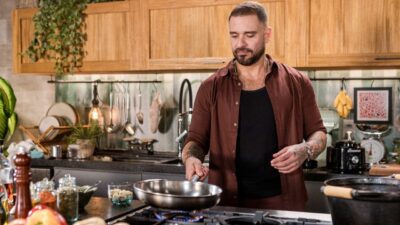 Cantor Diogo Nogueira cozinha em estúdio, veste camisa marrom aberta e regata preta, segura frigideira sobre o fogão, possui barba e tatuagens nos braços, fundo com armários de madeira clara e utensílios domésticos diversas em cozinha rústica moderna