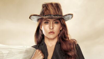 Angelique Boyer como Domênica Montero usa chapéu de couro bovino marrom, brincos de argola dourados e camisa preta com textura em cenário rústico