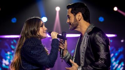 Emília (Dulce María) e Ángel (David Zepeda) cantam frente a frente com microfones em palco iluminado em cena da novela Penso em Ti