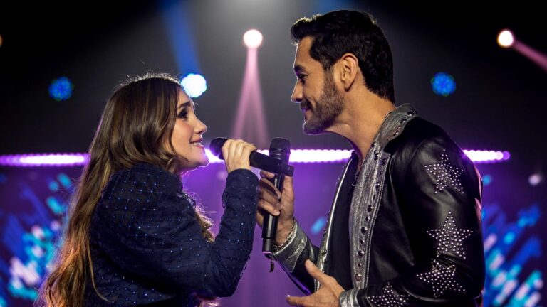 Emília (Dulce María) e Ángel (David Zepeda) cantam frente a frente com microfones em palco iluminado em cena da novela Penso em Ti