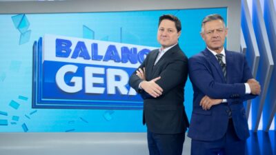 Eleandro Passaia e Diógenes Lucca posam de braços cruzados no estúdio do Balanço Geral SP, vestem ternos escuros, fundo com logotipo do programa em azul e branco