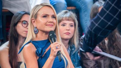 Apresentadora Eliana sorri e segura microfone preto, usa vestido azul com detalhe de flor no ombro e brincos grandes, plateia ao fundo no cenário do Altas Horas