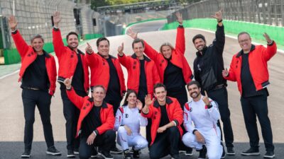 Equipe de transmissão da Fórmula 1 na Globo posa na pista de Interlagos, profissionais vestem jaquetas vermelhas e macacões brancos de corrida
