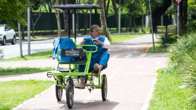 Chef Erick Jacquin pedala bicicleta verde de quatro rodas em ciclovia, vestindo colete e calça azuis, com placa do programa Pesadelo na Cozinha