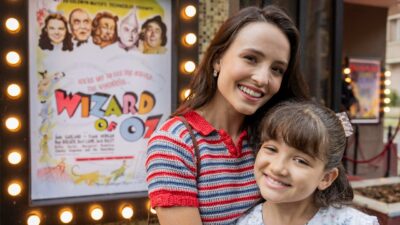 Larissa Manoela como Estela e Isabelly Carvalho como Anabela sorriem abraçadas na novela Êta Mundo Melhor!, ao lado de um cartaz de O Mágico de Oz