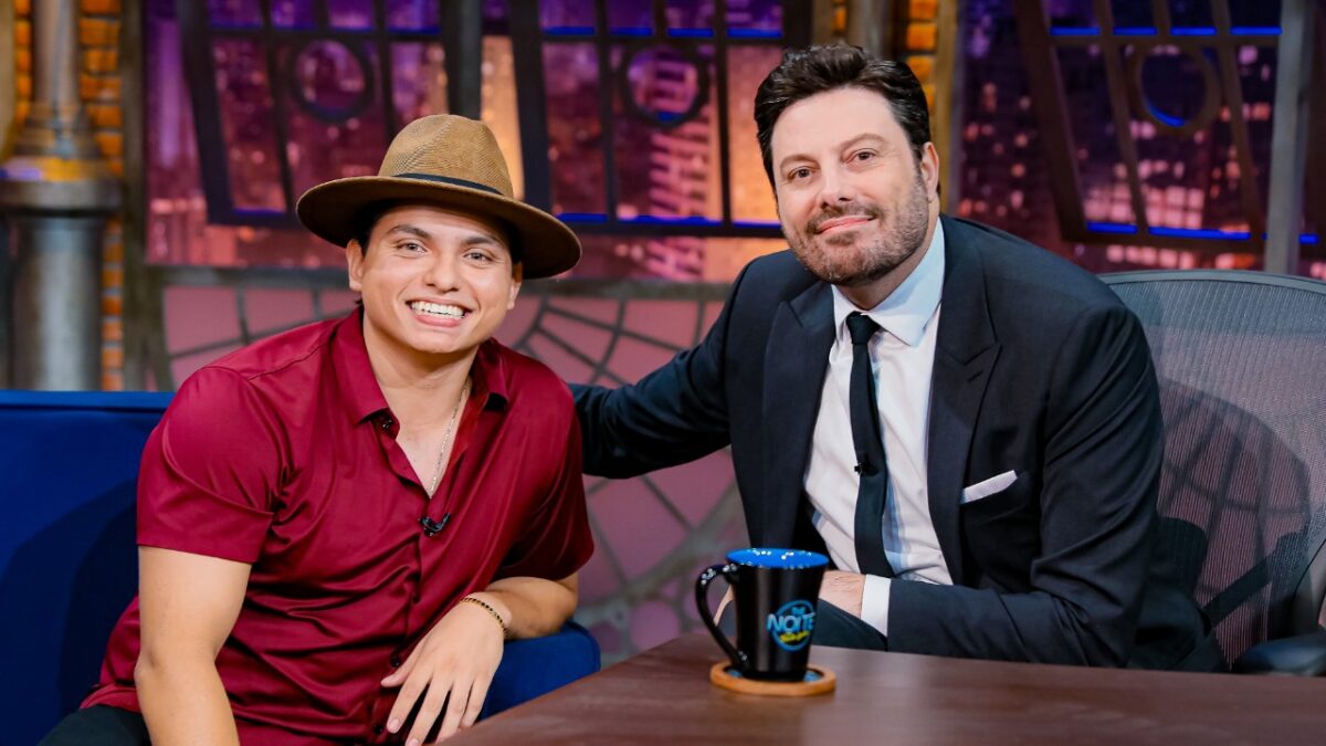 Everton da Silva, o Filho do Piseiro, de chapéu e camisa bordô, sorri ao lado de Danilo Gentili, de terno e gravata, no cenário do talk show The Noite