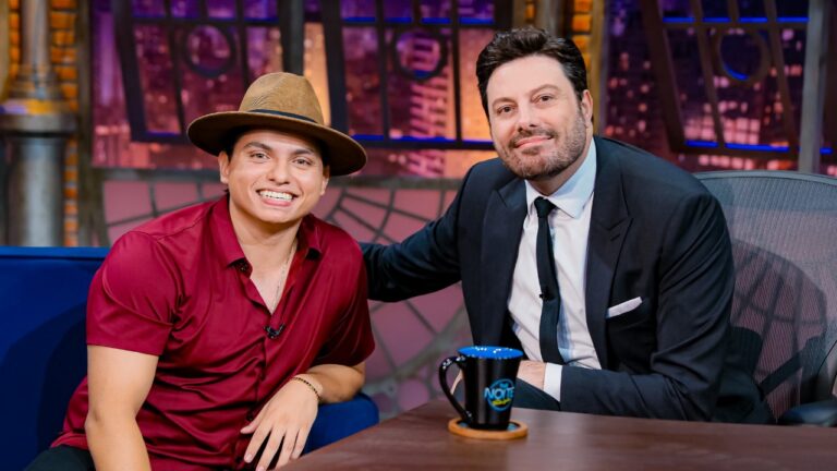 Everton da Silva, o Filho do Piseiro, de chapéu e camisa bordô, sorri ao lado de Danilo Gentili, de terno e gravata, no cenário do talk show The Noite