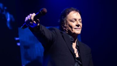 Cantor Fábio Jr. com olhos fechados, segura microfone em direção ao público, veste terno preto e camisa escura sob iluminação azul de palco em show
