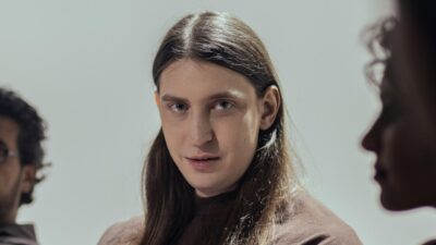 Felca com cabelo longo olha para o lado em cena de entrevista exibida no Fantástico com outras pessoas desfocadas ao fundo