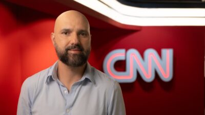 Fernando Rodrigues, de camisa social azul clara e barba, posa em frente ao logotipo luminoso da CNN Brasil em um cenário de fundo vermelho