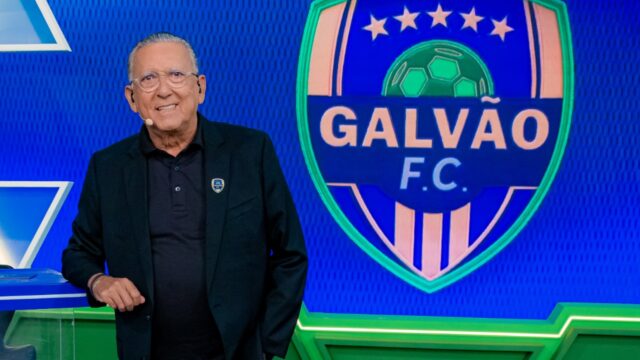 Galvão Bueno sorri no estúdio, vestindo blazer preto sobre camisa polo escura, posicionado ao lado do escudo do programa Galvão F.C. em um telão azul vibrante.