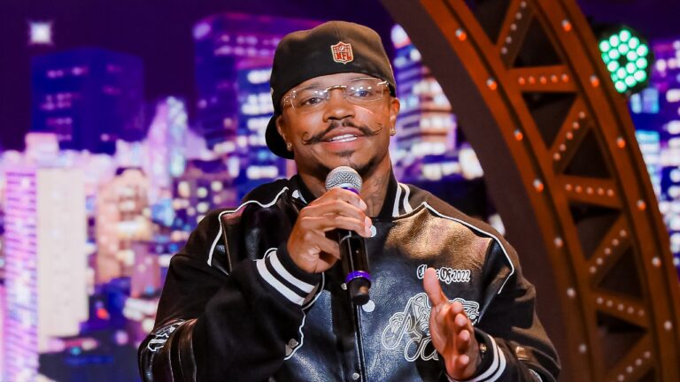 Cantor Gamadinho segura microfone, usa boné preto com logo da NFL e jaqueta bomber escura, exibe bigode estilizado no palco do programa The Noite clara