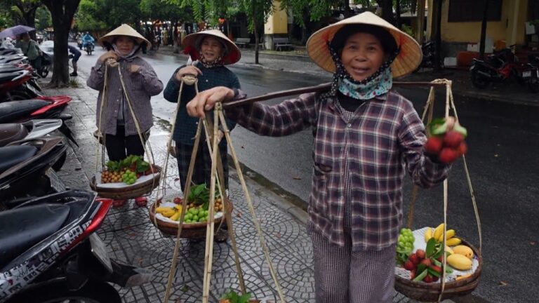 Vendedoras com chapéu cônico carregam cestos de frutas em vara de bambu em rua molhada no Vietnã durante gravação do Globo Repórter