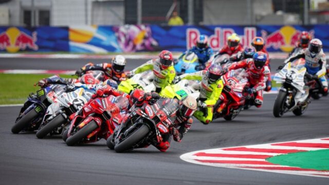 Pilotos de MotoGP inclinam motos coloridas em curva acentuada de pista asfáltica, competidores usam macacões e capacetes em alta velocidade no GP