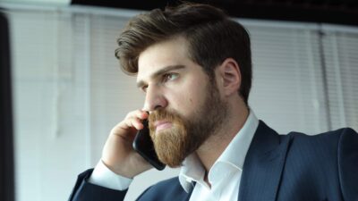 Berker Guven, interpretando Iskender em Chamas do Destino, aparece de perfil usando terno escuro e camisa branca enquanto fala ao celular em um escritório