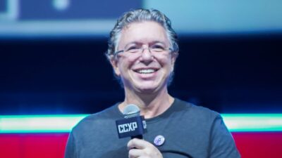 Boninho sorri e segura microfone da CCXP durante participação em painel no evento com palco iluminado ao fundo