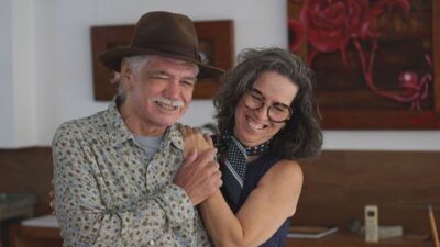 O ator Jackson Antunes, de chapéu marrom e camisa floral, sorri ao abraçar a esposa Cristiane Britto, que usa óculos e lenço azul de poá no pescoço
