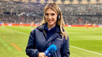Janice de Castro segura microfone com headset à beira do campo durante cobertura em estádio de futebol lotado ao fundo