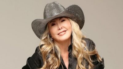 Cantora Jayne sorridente com cabelos loiros e ondulados, usando chapéu de cowboy cinza brilhante e camisa preta com detalhes em pedrarias no ombro