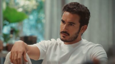 Ator Filipe Bragança como João Raul na novela Coração Acelerado, usa camiseta branca, tem barba e bigode, e observa atentamente em cenário interno