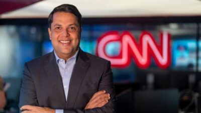 João Vitor Xavier sorri de braços cruzados em estúdio da CNN Brasil com logotipo vermelho ao fundo