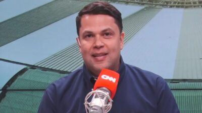 João Vítor Xavier fala ao microfone da CNN Brasil em estúdio com telão ao fundo durante transmissão de programa