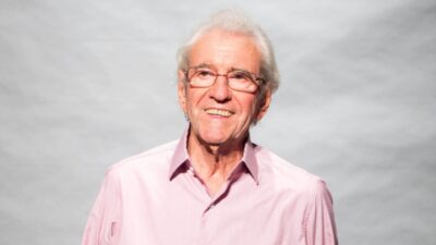 Juca de Oliveira sorri usando camisa rosa e óculos em retrato com fundo neutro cinza