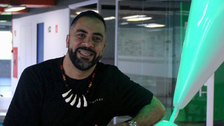 Jukanalha sorri na sede da RedeTV! com colar de dentes e camiseta preta apoiado em mesa com objeto iluminado ao lado