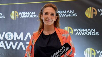 Juliana Algañaraz sorri segurando troféu no Woman Awards, veste quimono laranja com estampas de aves sobre blusa preta, fundo com logos do evento