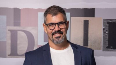 Juliano Cazarré posa sorrindo com blazer escuro e camiseta clara diante de painel com letras estilizadas ao fundo