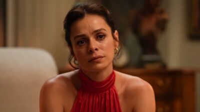 Atriz Ozge Ozpirincci, como Karsu na novela Coração de Mãe, encara a câmera com olhar melancólico, veste blusa vermelha de gola alta em sala de estar