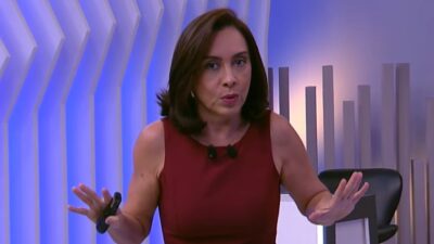 Jornalista Katiuzia Rios de blusa regata bordô gesticula com as mãos abertas no cenário do programa Brasil do Povo, com painéis brancos e luz azul