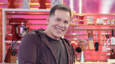 Leandro Hassum sorri usando jaqueta marrom sobre camiseta escura, posicionado à frente de cenário de cozinha com utensílios e fundo rosa iluminado clara