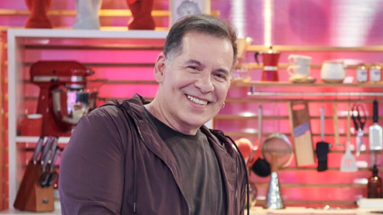Leandro Hassum sorri usando jaqueta marrom sobre camiseta escura, posicionado à frente de cenário de cozinha com utensílios e fundo rosa iluminado clara