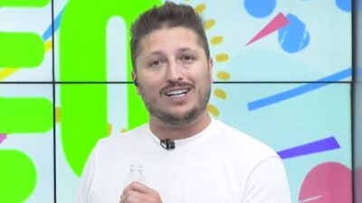 Léo Improta sorri e segura garrafa de água durante apresentação em estúdio colorido de programa da Band Campinas