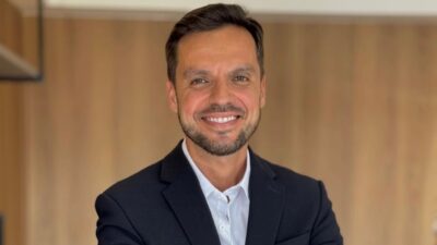 Léo Saballa sorri com braços cruzados usando terno escuro e camisa branca diante de parede de madeira em ambiente interno