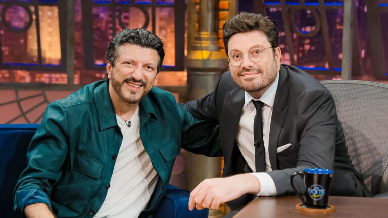 Lito Sousa, de jaqueta verde, e Danilo Gentili, de terno cinza e óculos, sorriem abraçados no sofá azul durante entrevista no programa The Noite