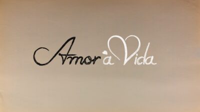 Logotipo da novela Amor à Vida com tipografia cursiva, palavra Amor em preto e Vida em branco com coração no topo da letra V, fundo bege claro clara
